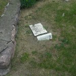 A rugged stone wall is adjacent to a broken concrete fragment resting on the grass, illustrating urban decay in nature.
